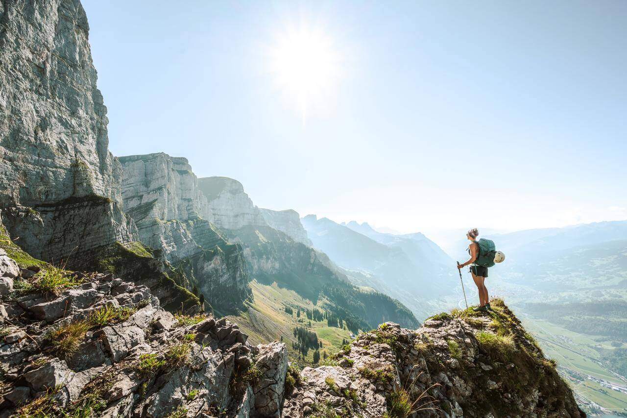 Eine Person steht auf einem felsigen Berggipfel und blickt in die Ferne. Der Himmel ist klar und die Sonne scheint hell. Im Hintergrund sind steile Bergwände und ein weites Tal sichtbar.