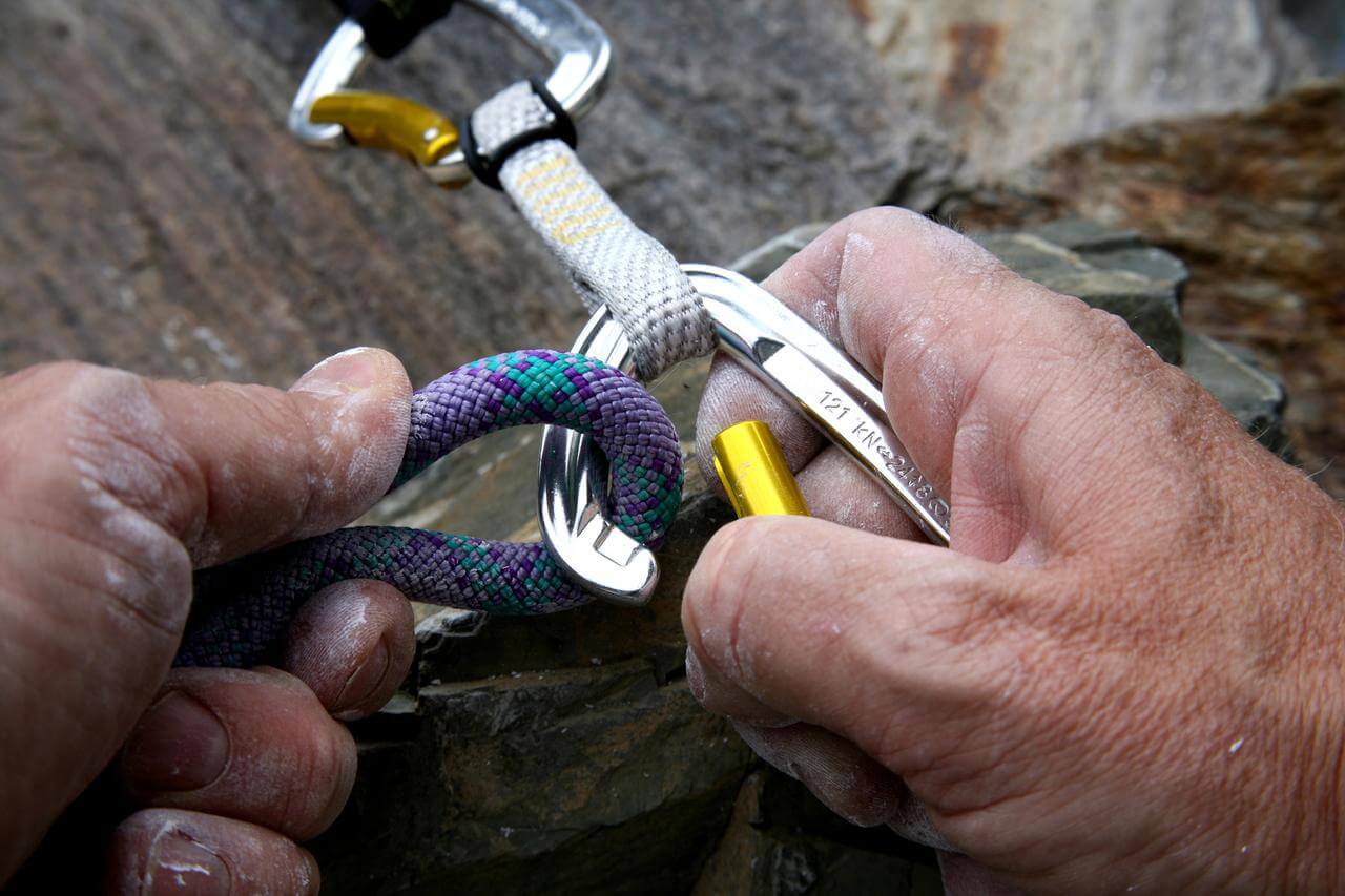 Nahaufnahme von zwei Händen, die Kletterausrüstung bedienen. Eine Hand hält ein buntes Kletterseil, während die andere einen Karabiner sichert. Die Ausrüstung scheint an einer Kletterwand im Freien verwendet zu werden.