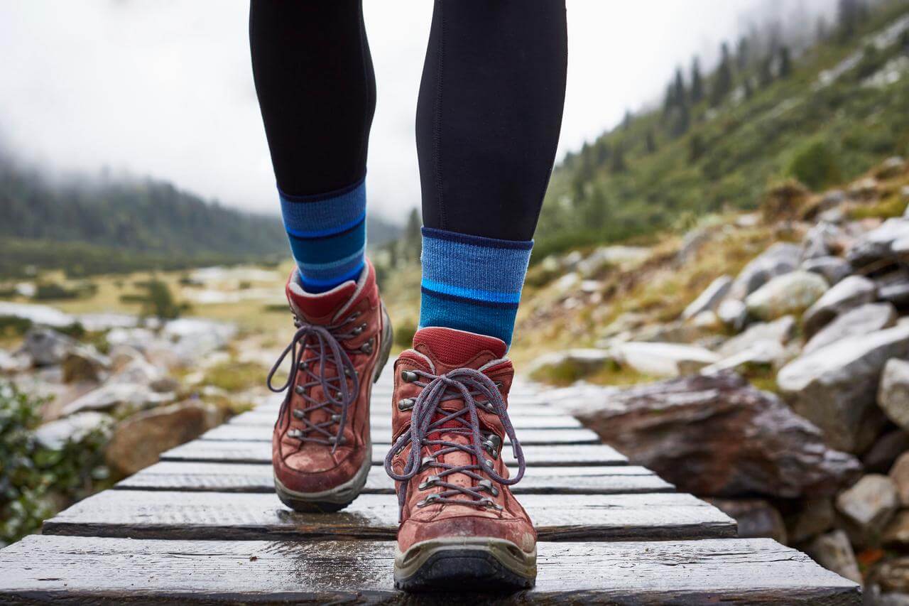 Nahaufnahme von einem Paar wandernden Füßen in roten Wanderschuhen und blauen Socken, die auf einem Holzsteg in einer bergigen Landschaft spazieren. Im Hintergrund sind unscharf Grünflächen und Felsen zu erkennen, die von einer wolkigen, nebligen Atmosphäre umgeben sind.
