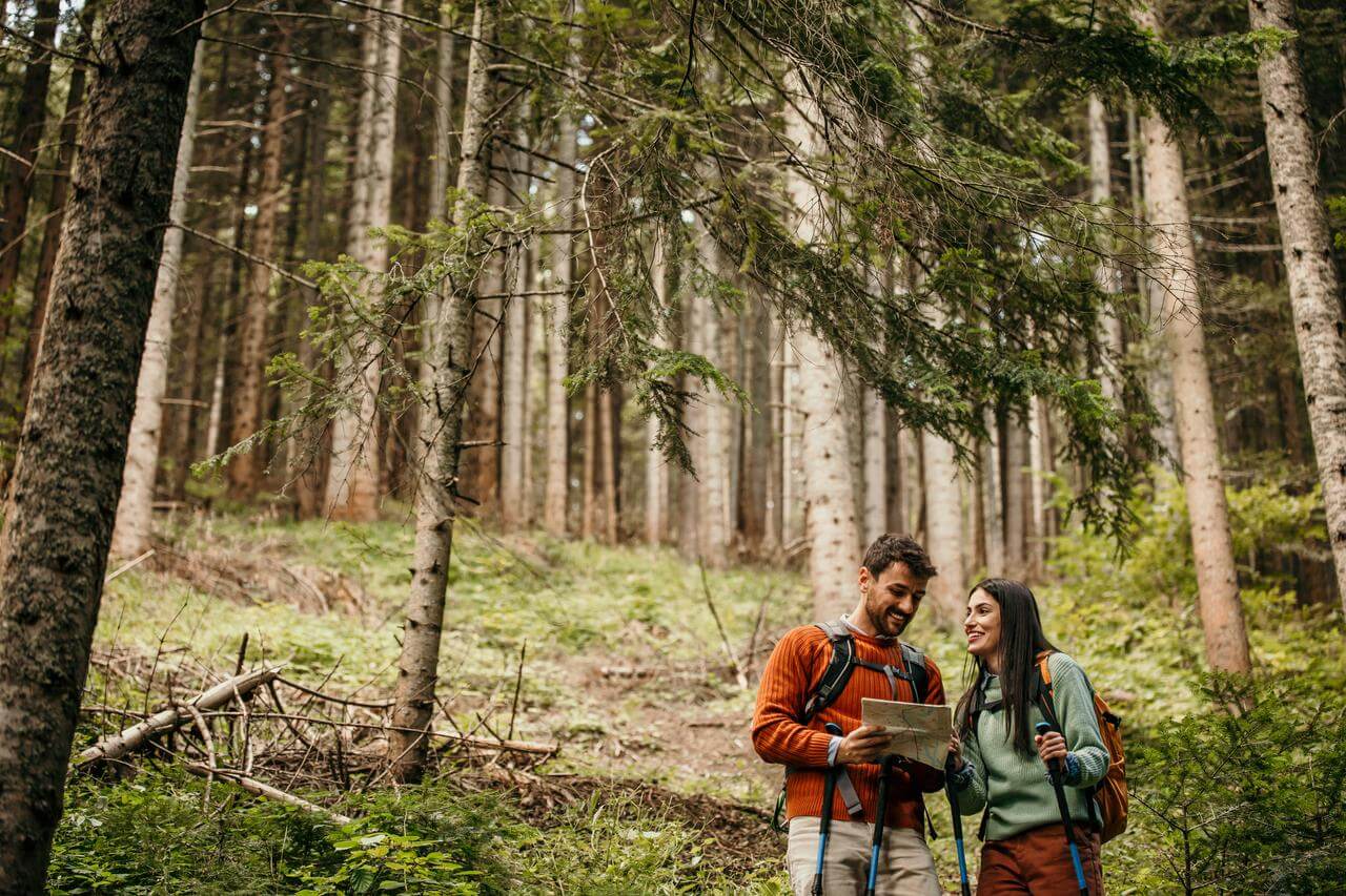 Ein Mann und eine Frau wandern im Wald und betrachten eine Karte. Beide tragen Rucksäcke und Outdoor-Kleidung. Die Umgebung ist voller Bäume und grünem Laub.